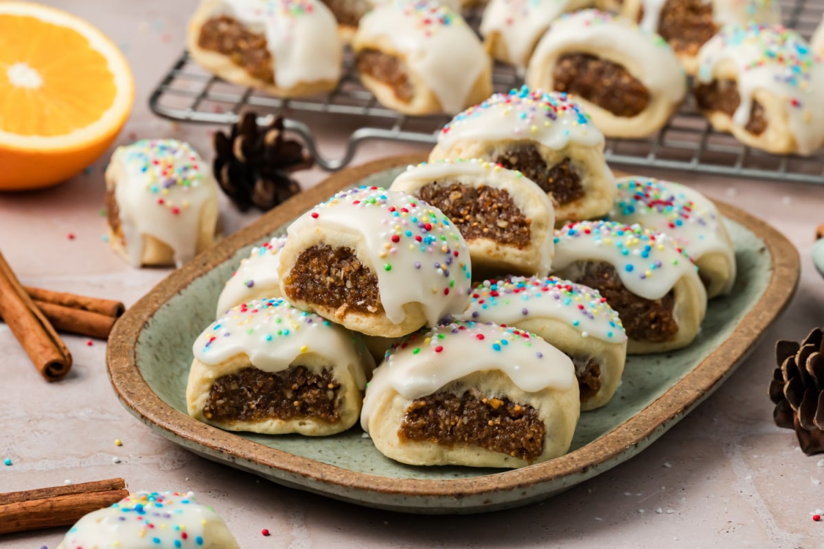 Cuccidati Cookies with Moist Fig Filling and Vanilla Glaze Italian fig cookies arranged on a ceramic platter, showing the moist fig and walnut filling inside buttery pastry topped with vanilla icing and rainbow sprinkles.