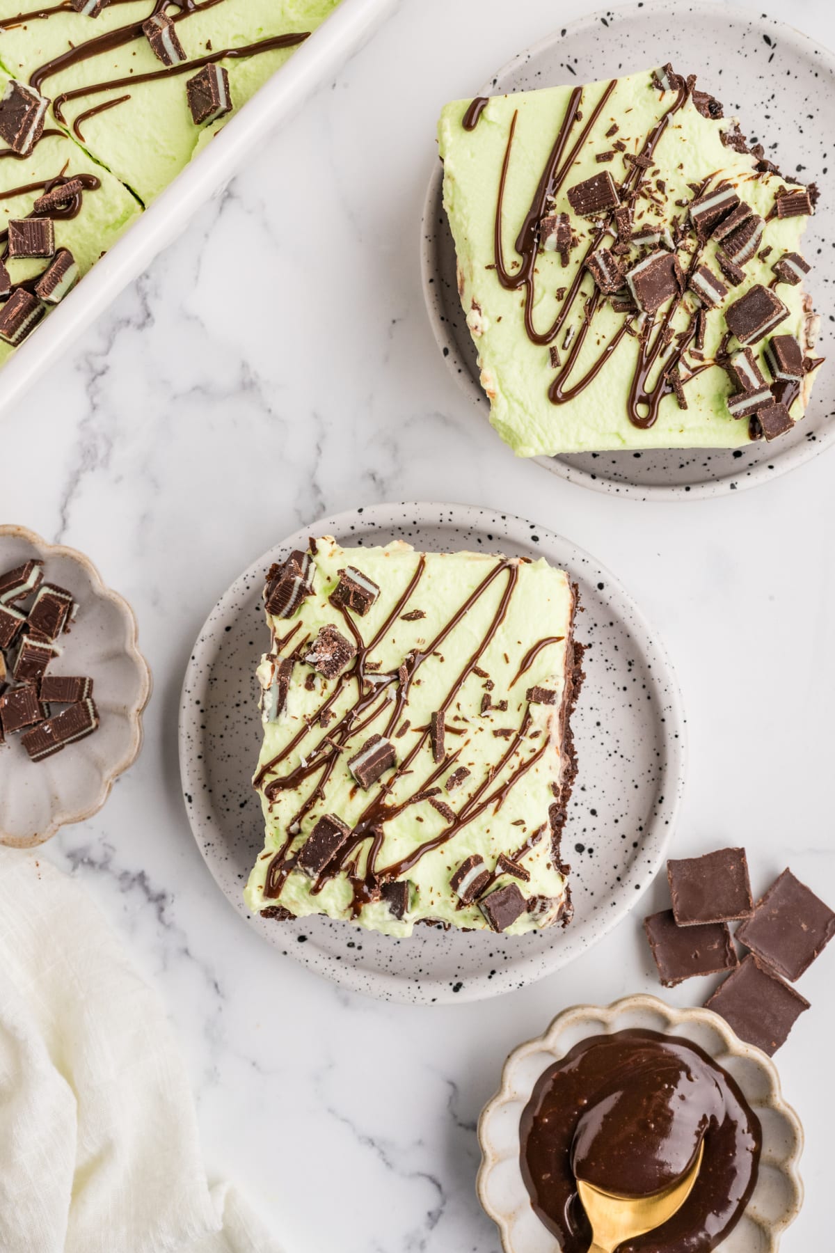 Mint Chocolate Poke Cake with Andes Mint Topping Two slices of mint chocolate poke cake on speckled plates with pale green whipped cream, chopped Andes mints and hot fudge drizzle.