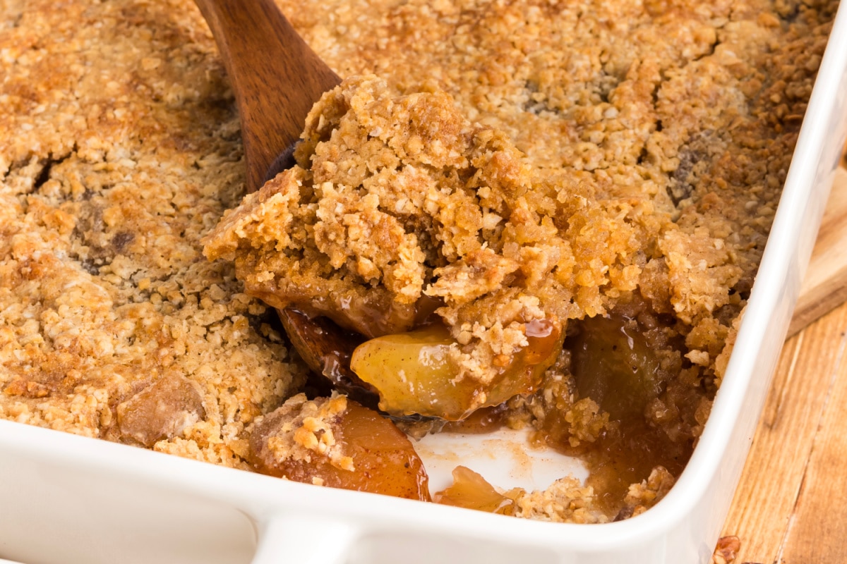 A spatula lifting oatmeal cookie apple crisp dump cake from a baking dish, showing the cracked cookie crust and cinnamon apple filling.
