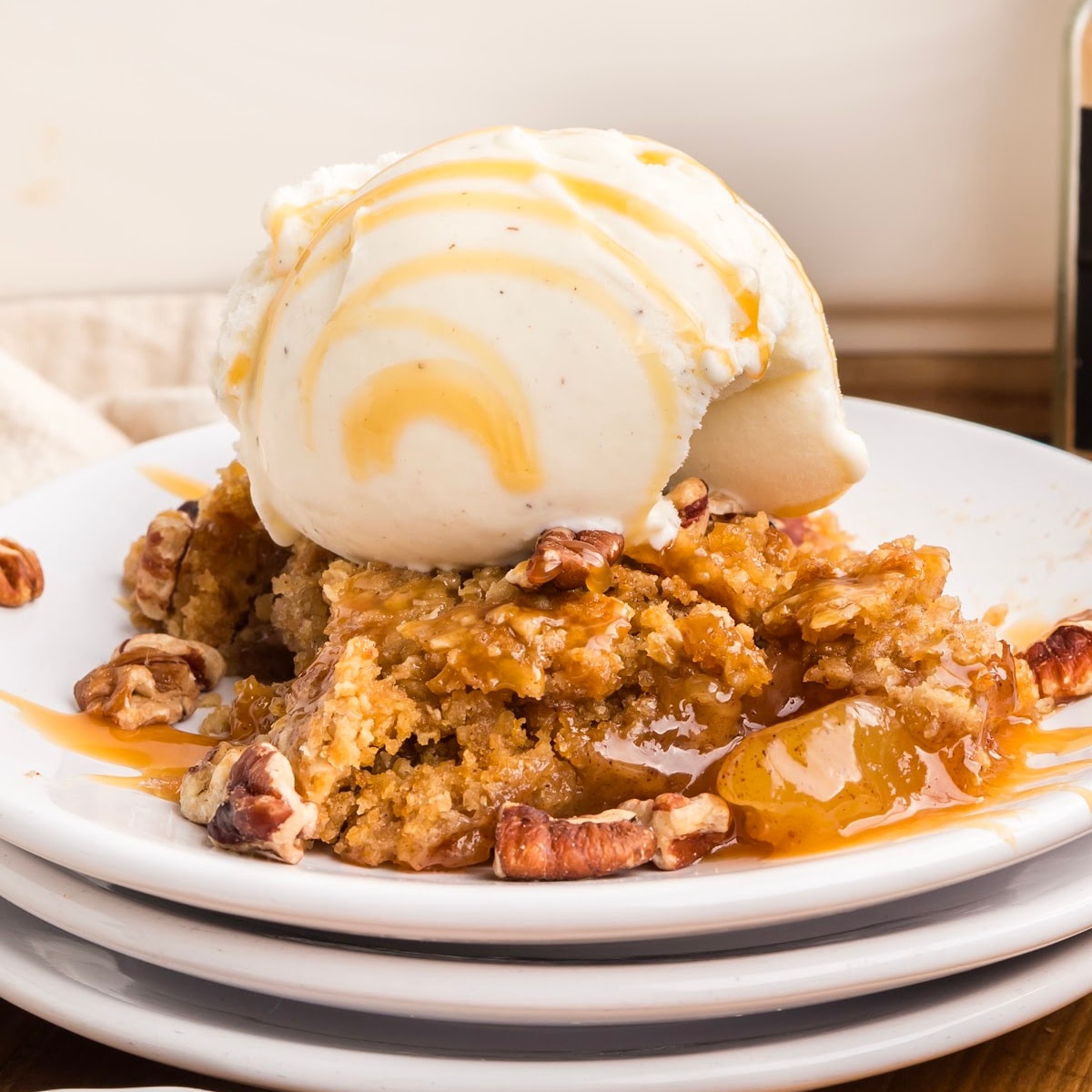 Oatmeal cookie apple crisp dump cake served on a white plate with a golden cookie topping, cinnamon apples, caramel sauce, pecans, and vanilla ice cream.