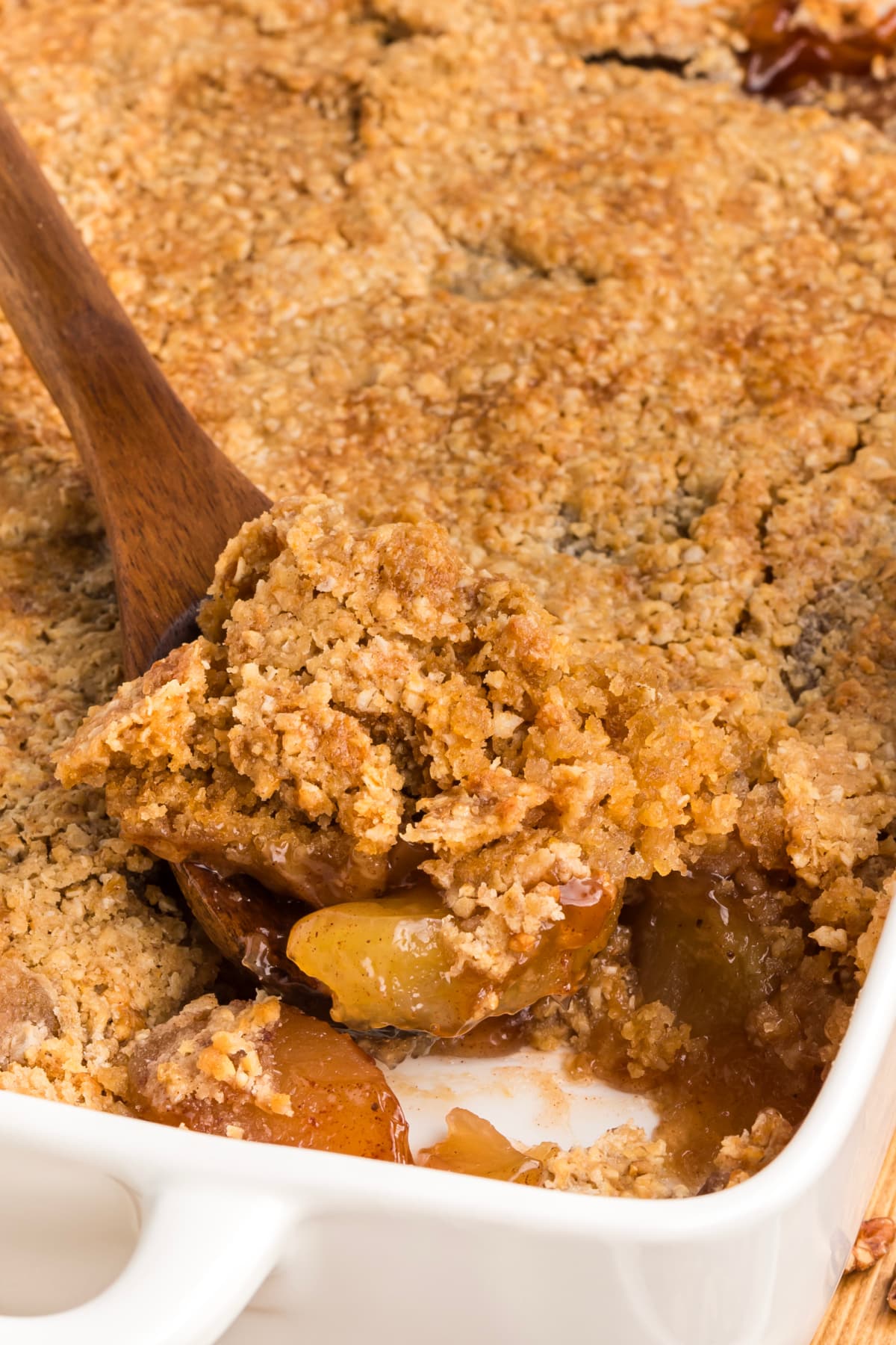 Wooden spatula lifting a serving of apple crisp with a cracked oatmeal cookie crust and soft cinnamon apple filling.