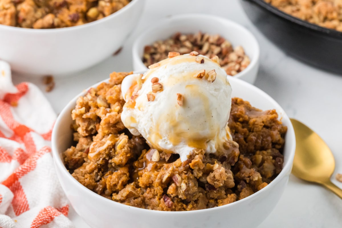 Bowl of pumpkin crisp with buttery oat and pecan topping, vanilla ice cream, and caramel drizzle.