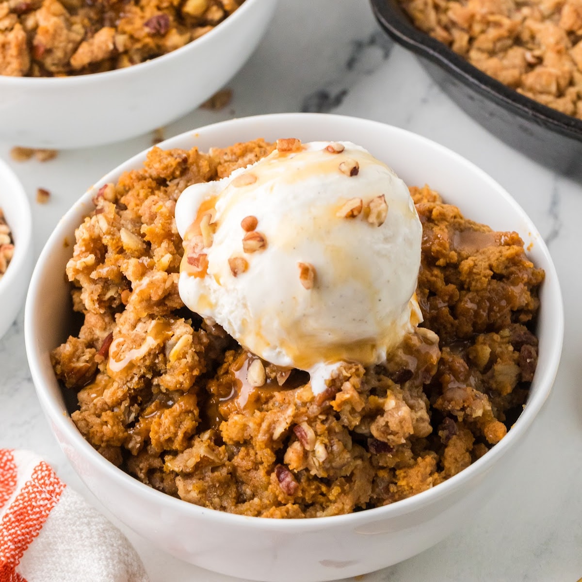 Pumpkin crisp with buttery oat and pecan topping, vanilla ice cream, and caramel sauce served in a white bowl.
