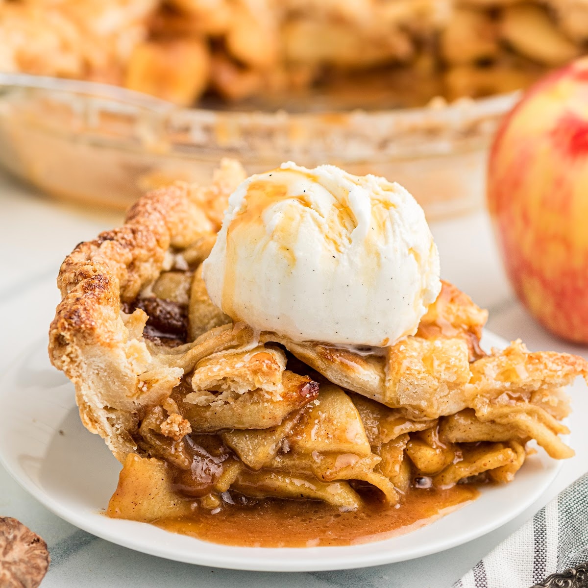 Homemade salted caramel apple pie slice topped with vanilla ice cream, showing thinly sliced apples coated in caramel beneath a golden lattice crust.