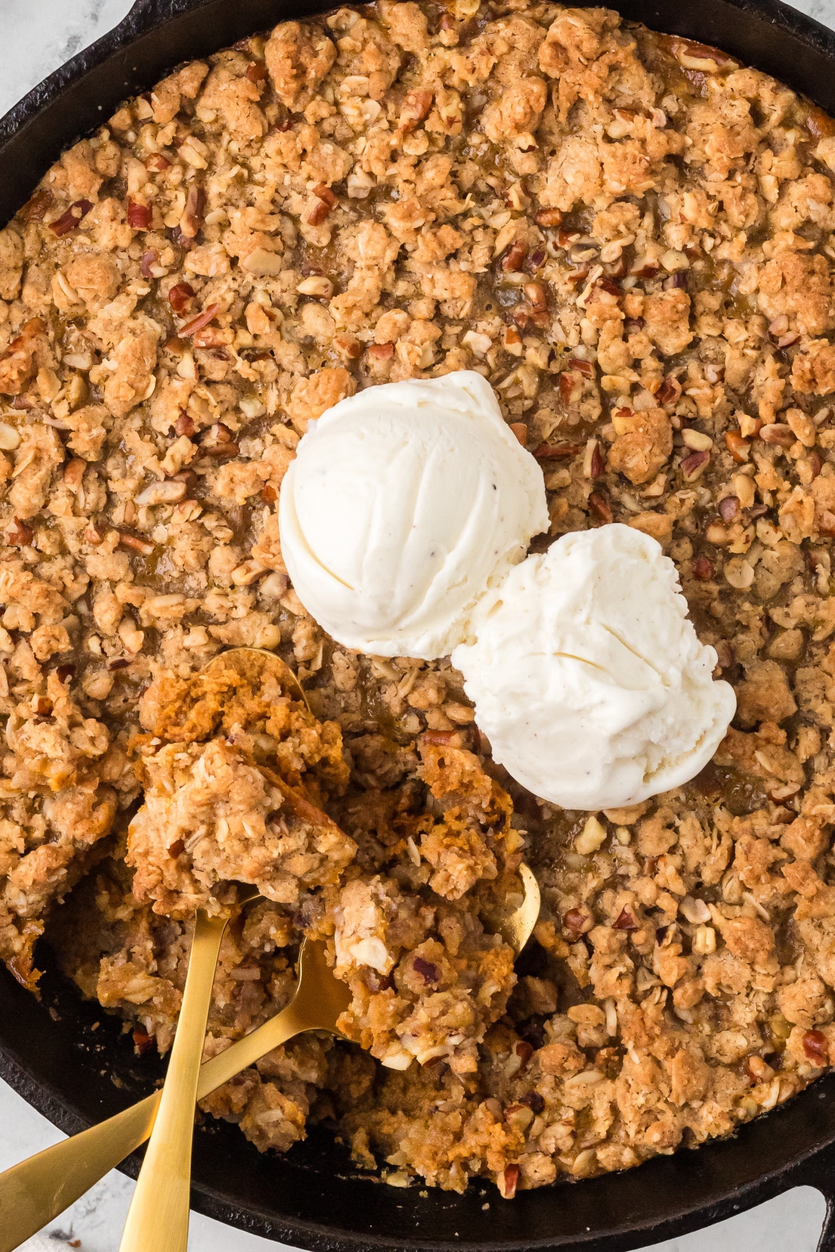 Cast-iron skillet filled with pumpkin custard and oat pecan topping, topped with two scoops of vanilla ice cream and partially served with gold spoons.