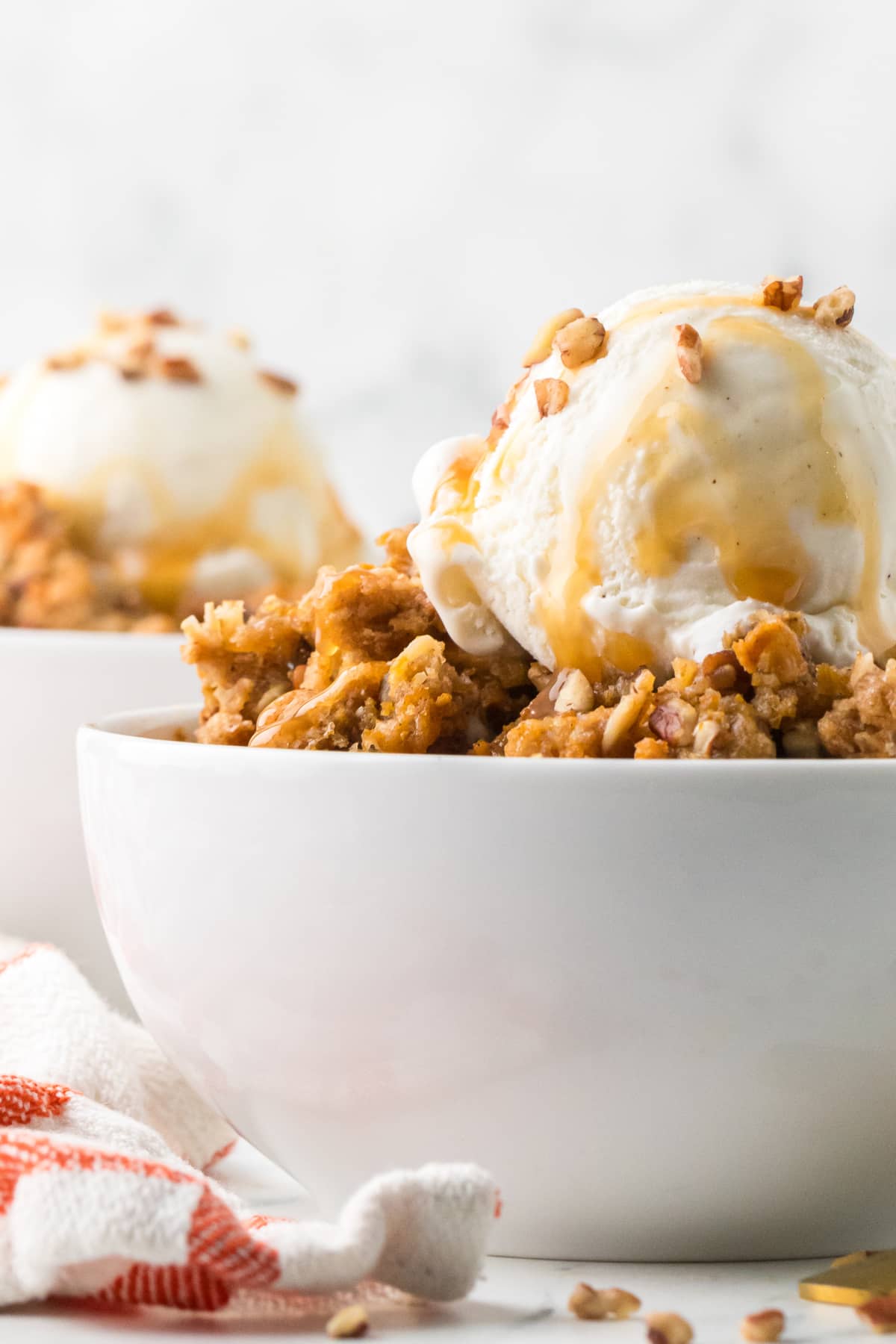 Side view of a bowl of spiced pumpkin custard with oat and pecan topping, vanilla ice cream, and caramel sauce.